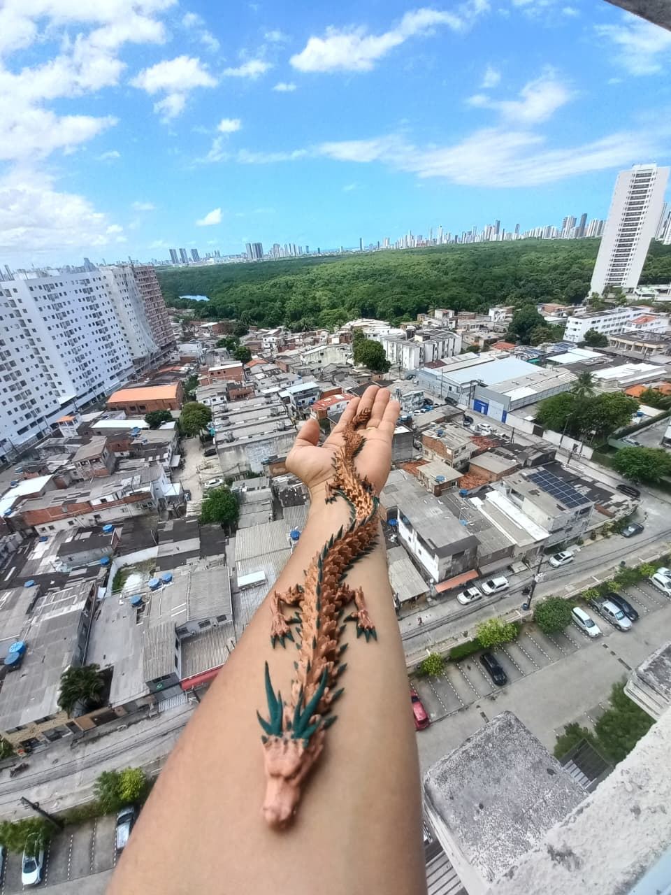 Dragão articulado impresso em 3D sobre o braço, com Recife ao fundo